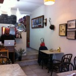 Imagem de uma pequena cafeteria, com algumas mesas, quadros na parede de utensilios de cozinha antigos, um balcão ao fundo com uma maquina de café espresso. O barista está sentado em frente ao balcão, usando um computador.
