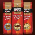 imagem das embalagens de cafés aromatizados do café do ponto. Trata-se de uma embalagem de alumínio retangular, com o da empresa e o título "Gourmet Café" e um rótulo circular especificando o sabor. Na foto, há três opções: Amendoas Torradas, Chocolate com trufas e Creme Irlandês.