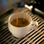 Imagem de uma xícara de café sob a máquina de café espresso, com o líquido caindo na xícara.