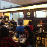 Imagem do salão de café, com várias mesas redondas e cadeiras. Há várias pessoas sentadas e pode-se ver o balcão ao fundo e espelhos nas paredes atrás do balcão e das mesas.
