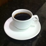 Imagem de uma xícara de café, sobre o pires. Numa mesa de marmore preta, em contraste com a xícara branca.
