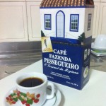 Imagem de uma xícara de café pequena, sobre um pires. Ambos brancos, com desenho de morangos e folhas. O pires tem também uma linha verde que circula toda sua extensão. Em segundo plano, a embalagem do café, fina e retangular, com topo fechado em forma de triangulo, onde tem o desenho de telhas, imitando o telhado de uma casa. Na embalagem, há o desenho de uma casa antiga branca com detalhes das portas e janela azuis. E, mais baixo, um fundo azul escuro e, em letras brancas, os dizeres: Café Fazenda Pessegueiro, O Gourmet da Mogiana. Café Torrado e Moído - Ground Roasted Coffee - peso líquido: 250g. Os produtos estão sob uma tábua branca, em cima de uma pia de alumínio. Ao fundo, pode-se ver a parede de azulejos beje e, em ultimo plano, outra xícara de café, vazia e virada pra baixo.