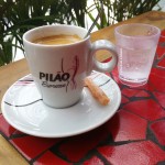 Foto: Lak Lobato - imagem de uma xícara de café branca, com o logo da marca escrito "Pilão Espresso" sobre um pires da mesma cor. Ao lado da xícara, um copo de 50ml de água com gás. Ambos sobre uma mesa de mosaico de ladrilho vermelho com borda de madeira. Em ultimo plano, imagem de folhas de alguma planta.